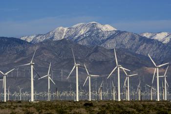 Farm of wind turbines in the foreground of a snowcapped mountain.