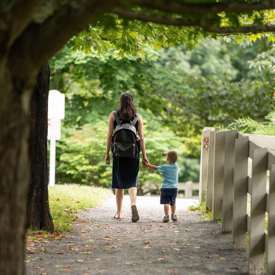 Young woman holding hands with small child walking down a path in a park.