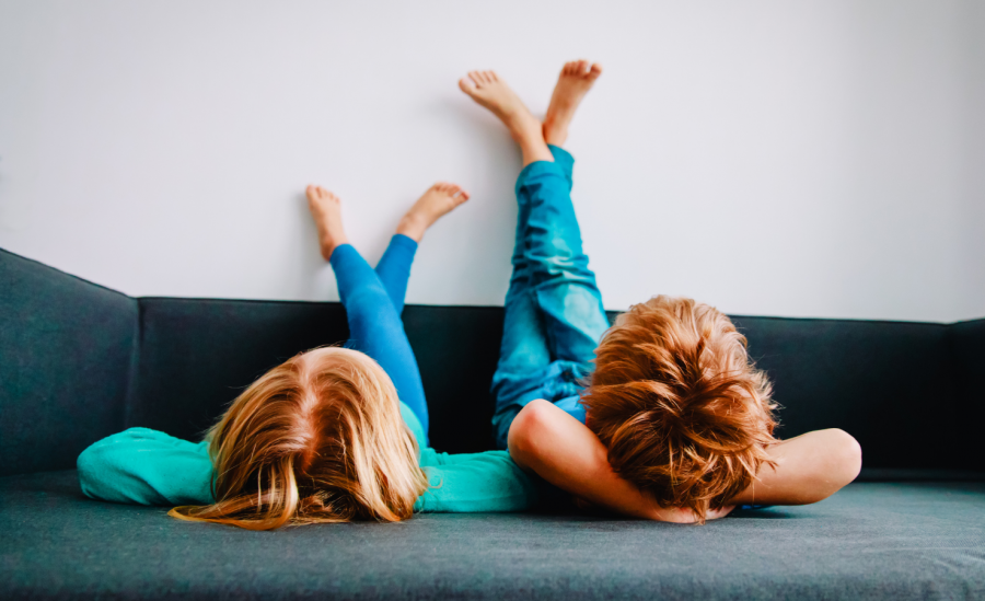 Two young kids laying comfortably on couch with arms folded and legs against wall.