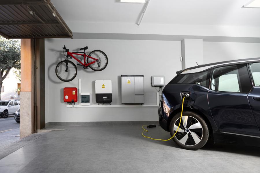 a car parked in a garage and plugged in to charge. a bike hangs on the wall by the garage door and electric boxes hang on the wall below the bike. 
