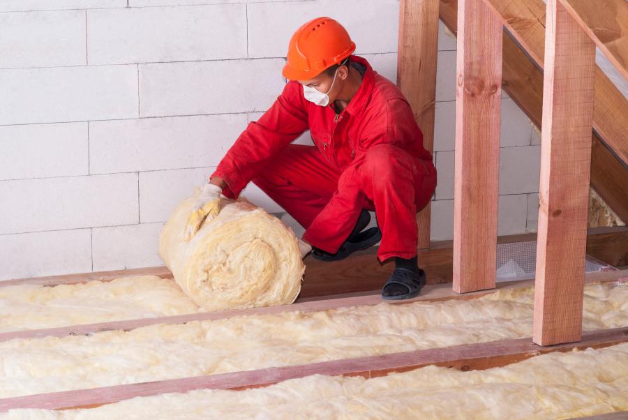 a person in a red jumpsuit with a facemask squatting down to roll insulation between ceiling joists