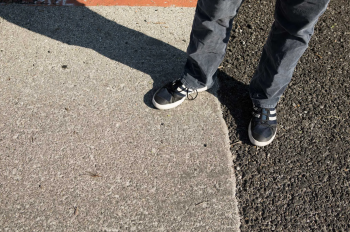 looking down at the ground, from the knees down there are two feet wearing sneakers with one on concrete and another on pavement