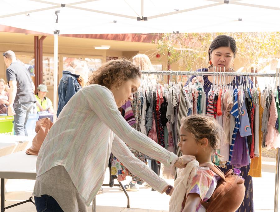 an adult puts a dress up to a child while another person looks through a rack of clothes in the background under a tent at a festival