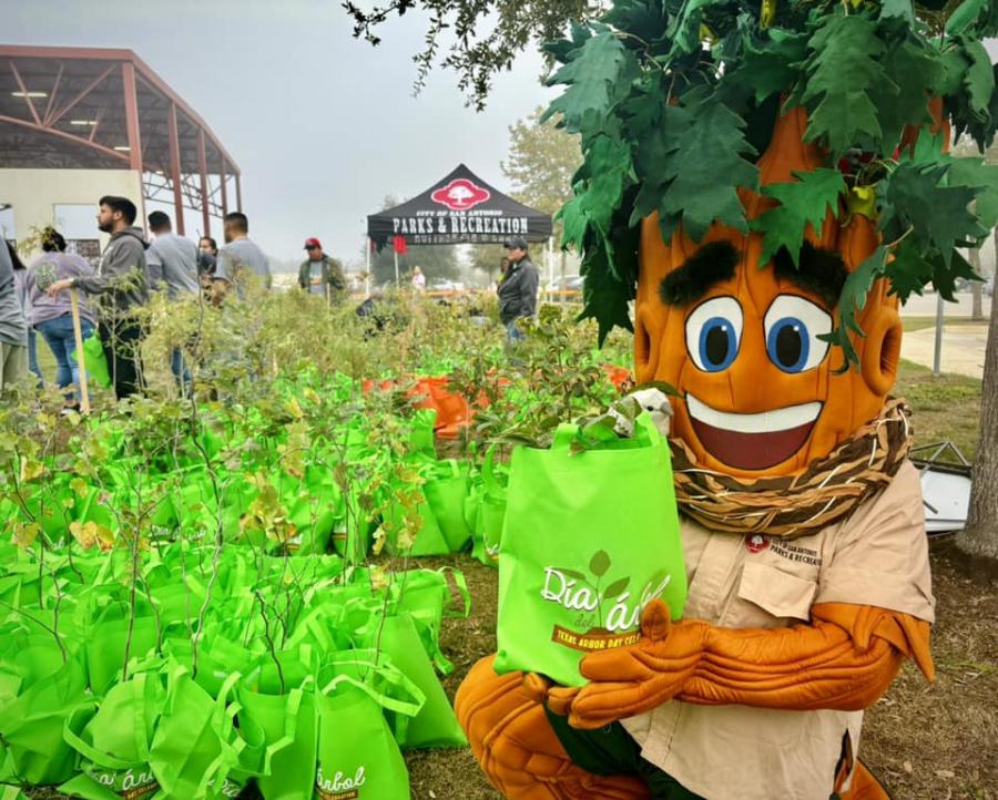 a tree mascot holding a bag with a plantable tree in front of many more bags!
