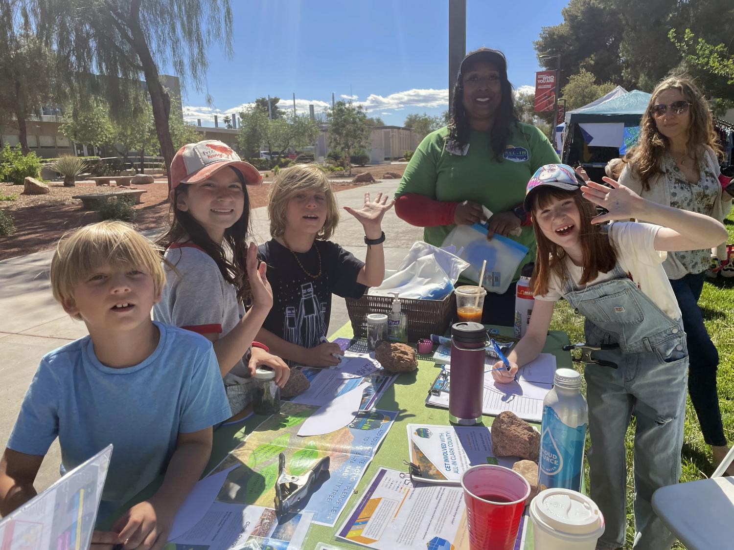 mostly kids around a table at an educational event. some are waving toward the camera excitedly