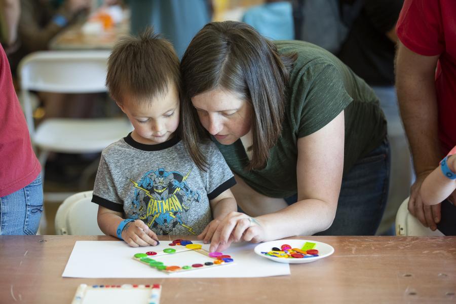 An adult leaning down and helping a child with an art project at a table