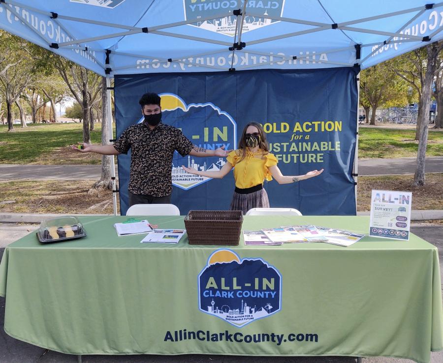 <p>two people standing under a tent at a table with arms outstretched welcomingly and masks over their mouths</p>