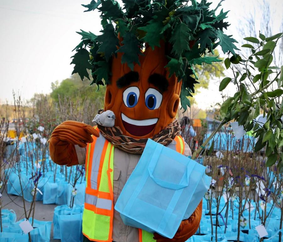 A person dressed as a tree mascot holds a reusable bag with a small tree in it