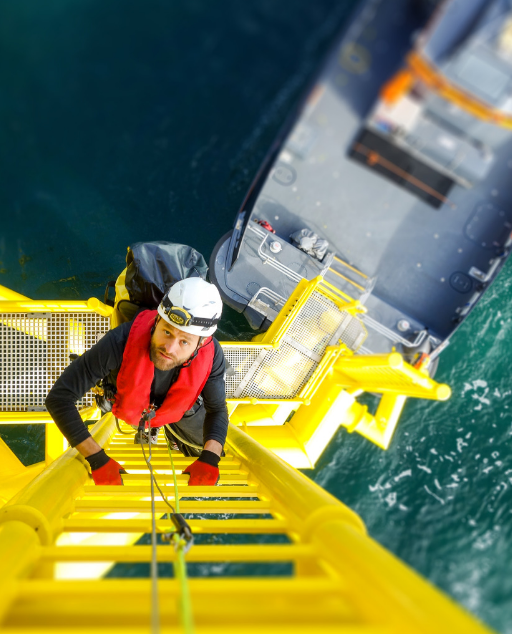 Man wearing safety gear climbing up a tall yellow ladder over a barge boat on the ocean.