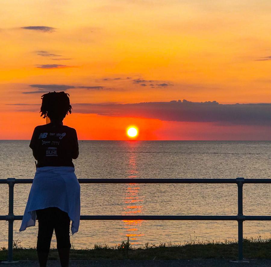 Silhouette of a young girl standing in front of a fence with a sunset over the ocean in the background.
