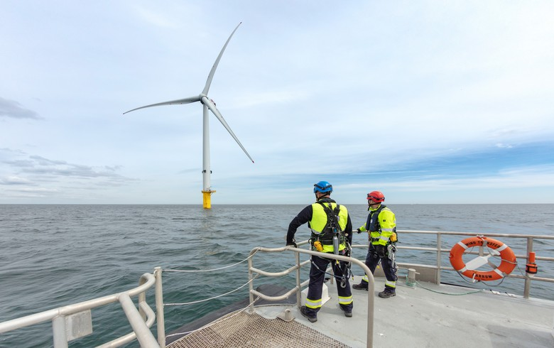 Two people in black and yellow safety outfits stand on the bow of a boat looking at an offshore wind turbine.