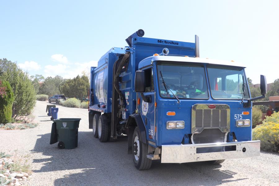 Photo of a blue recycling truck next to waste bins on the side of the road.