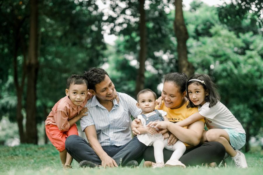 Smiling family of five sits cross legged on the grass of a forested park.