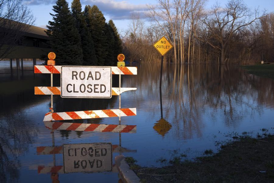 Road closed sign hangs on a traffic blockage submerged in water in a flooded rural street.
