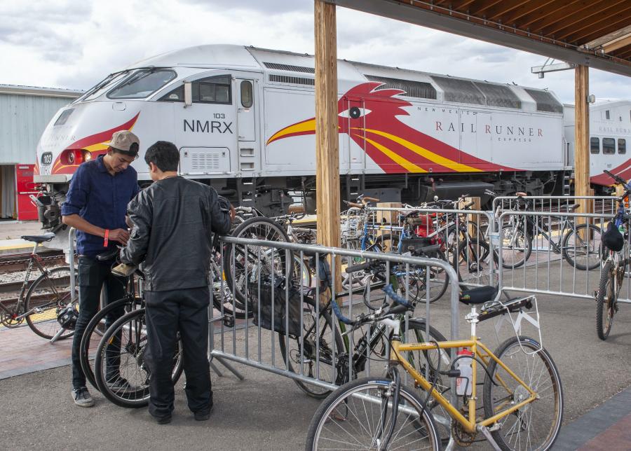 Photo of the Santa Fe train station with a Rail Runner Train in the background. In the foreground, two people stand next to bike racks in front of the train.